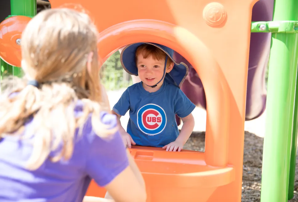  A toddler in a blue sunhat and blue Cubs shirt looking at a teacher through the window of an orange outdoor playscape