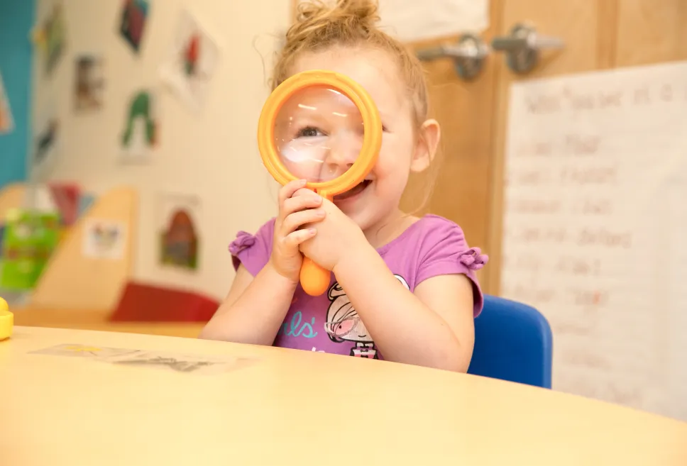A smiling toddler in a purple shirt looking through a magnifying glass