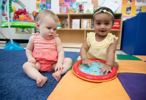 Two babies sitting on a yellow, blue and purple play mat 