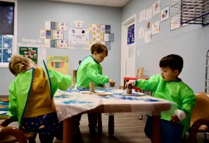 Three toddlers in green smocks working on an art project using paper plates and cardboard tubes