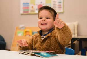 Child in a fuzzy brown sweater pointing and smiling while looking at a children's book