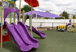 Outdoor play area for active learning at Little Sprouts in Medford, MA featuring a playscape with side-by-side purple slides and a shaded sandbox 