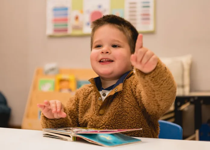 Child in a fuzzy brown sweater pointing and smiling while looking at a children's book