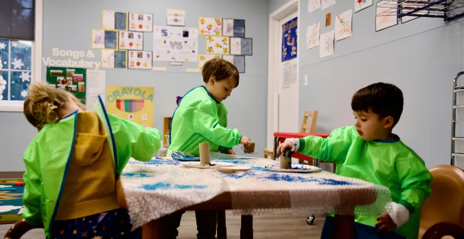Three toddlers in green smocks working on an art project using paper plates and cardboard tubes