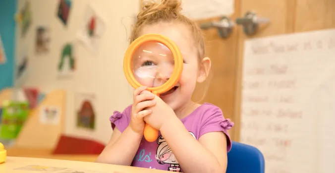 A smiling toddler in a purple shirt looking through a magnifying glass